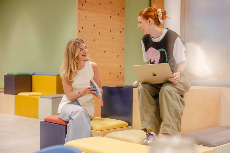 Two women sat on different stool levels, talking and smiling and looking at a book.